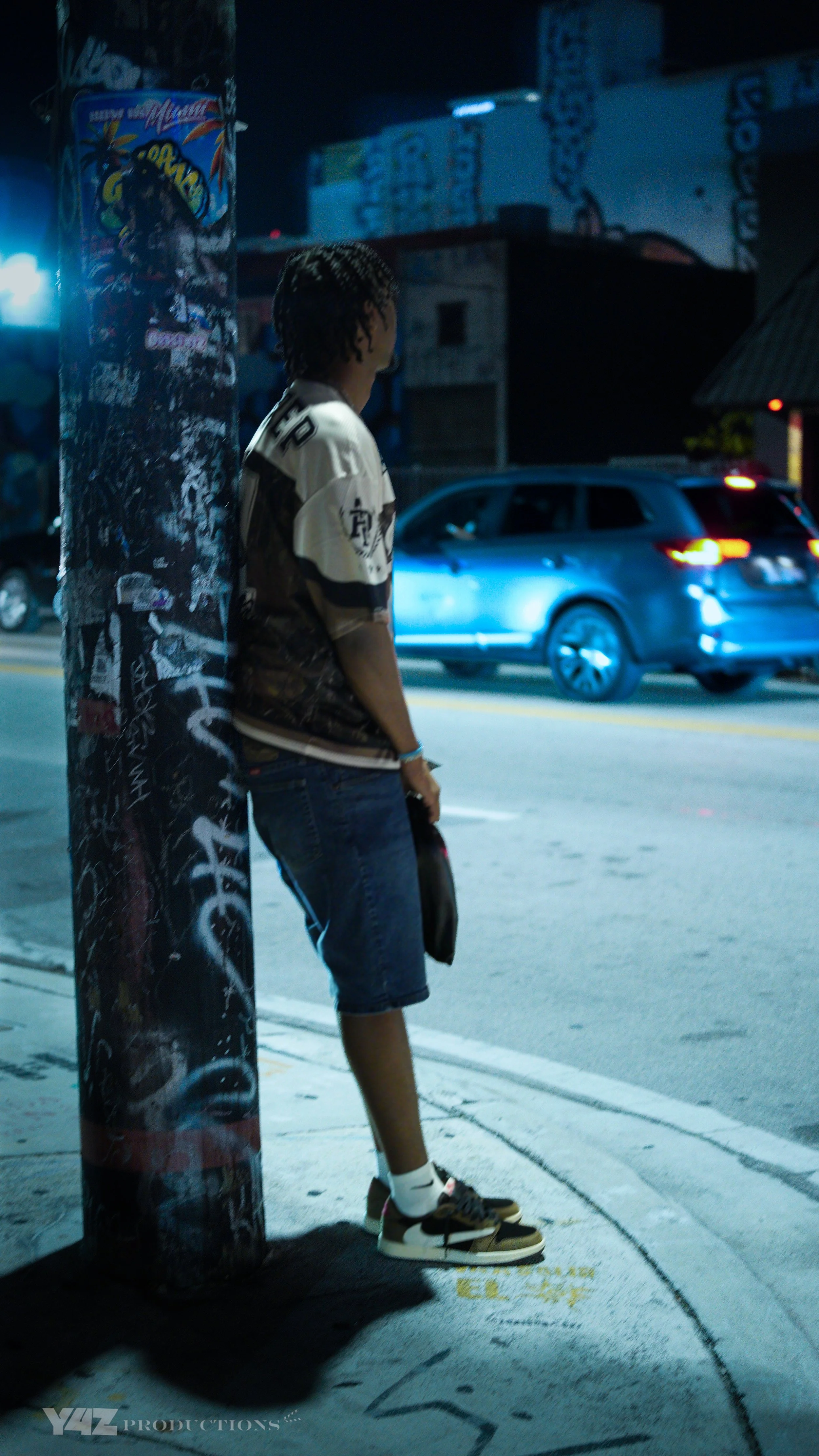 A young man with braided hair standing on a city sidewalk at night, leaning against a graffiti-covered pole, holding a face mask and wearing a graphic T-shirt, shorts, and sneakers. Cars are passing on the street behind him, and there is signage and buildings in the background.