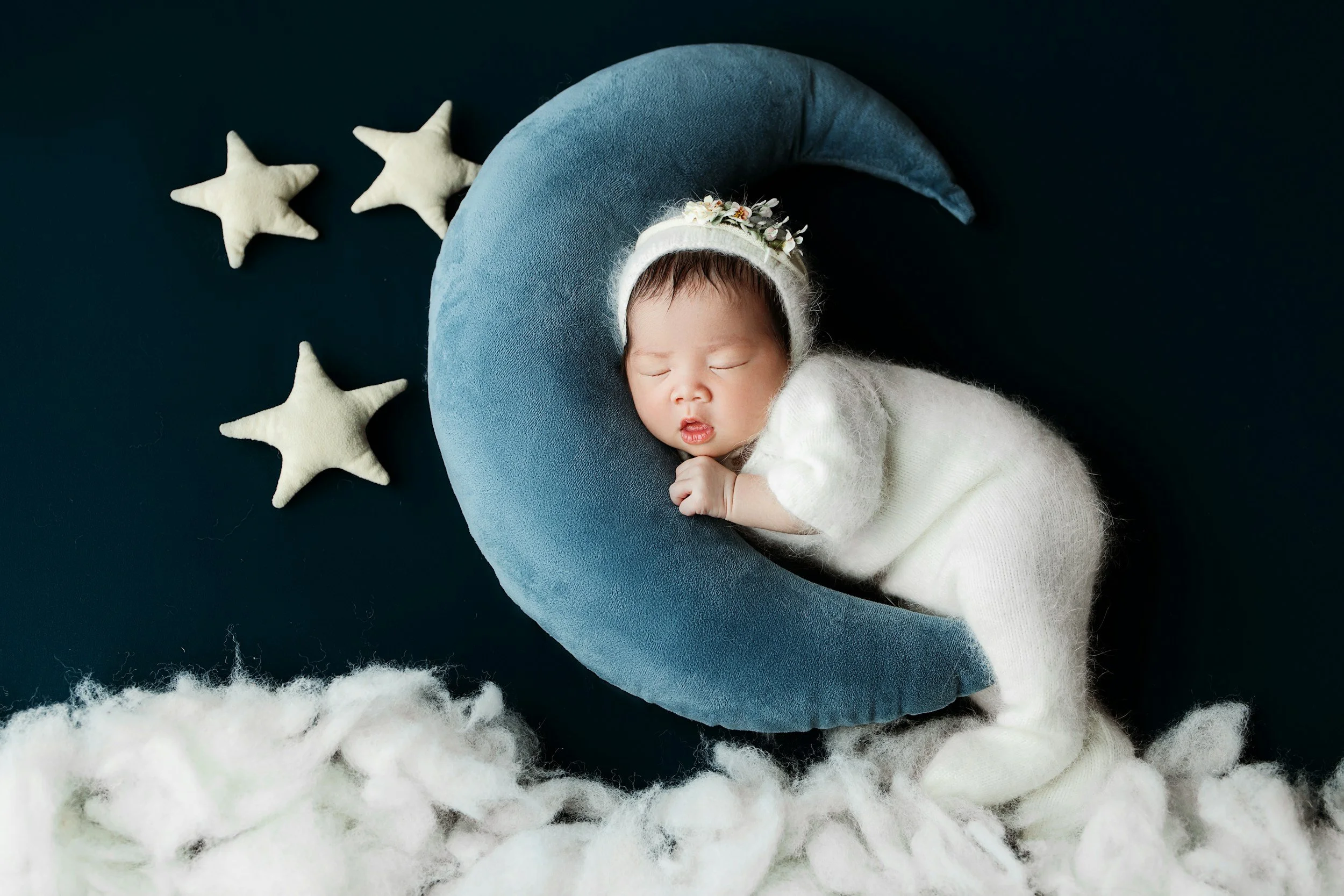 Sleeping baby in white outfit with a floral headband, resting on a blue crescent moon cushion surrounded by star-shaped plush toys and fluffy white material.