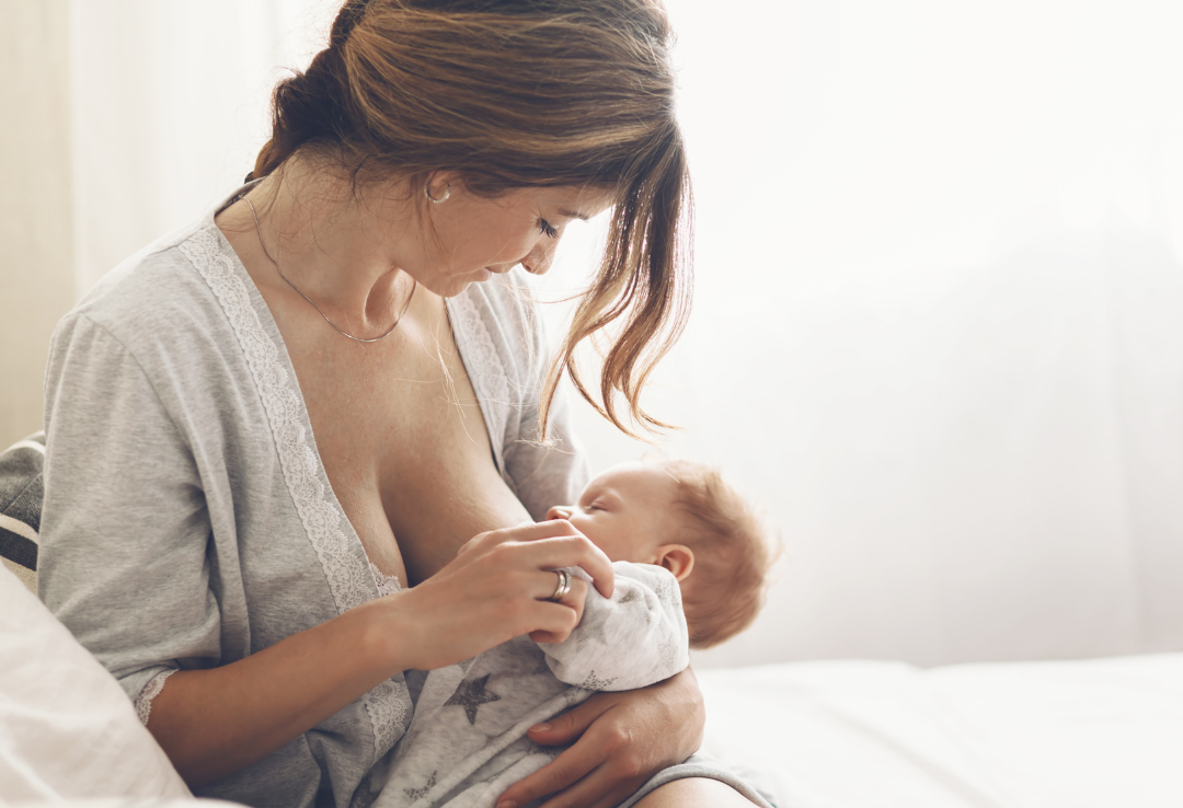 A woman with brown hair holding a baby at home, breastfeeding the baby while sitting on a bed.