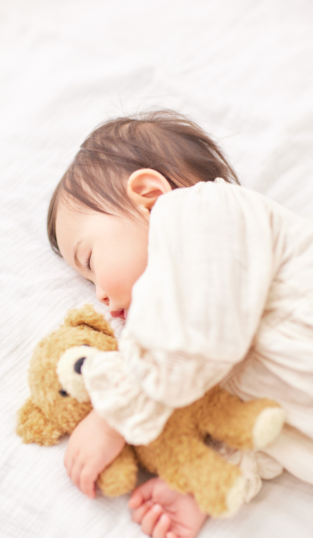 A young child sleeping peacefully on a white bed, hugging a brown teddy bear.