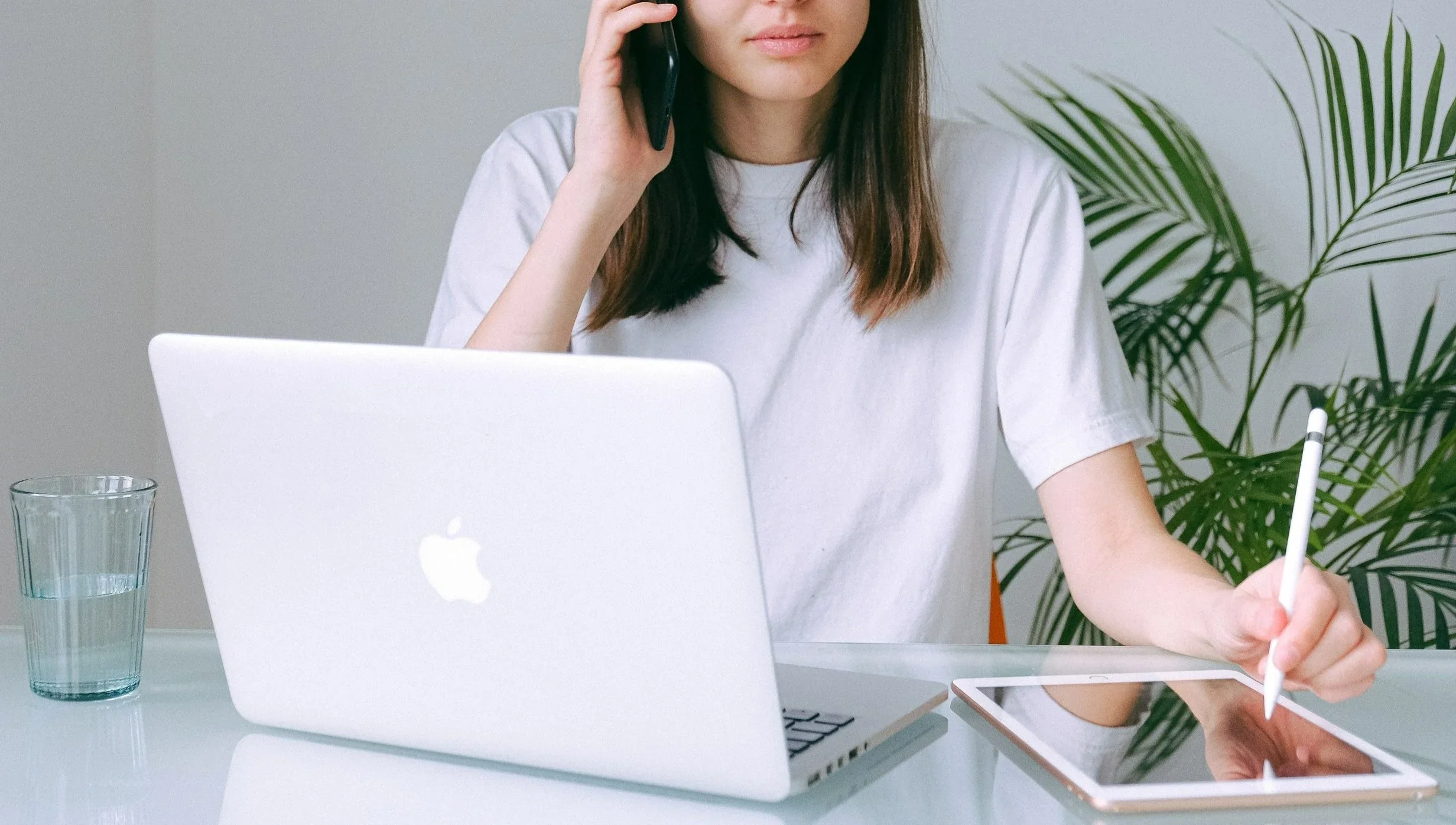 Woman sitting at a desk, talking on a phone, using a stylus on a tablet, with a silver MacBook and a glass of water on the desk, and a large plant in the background.