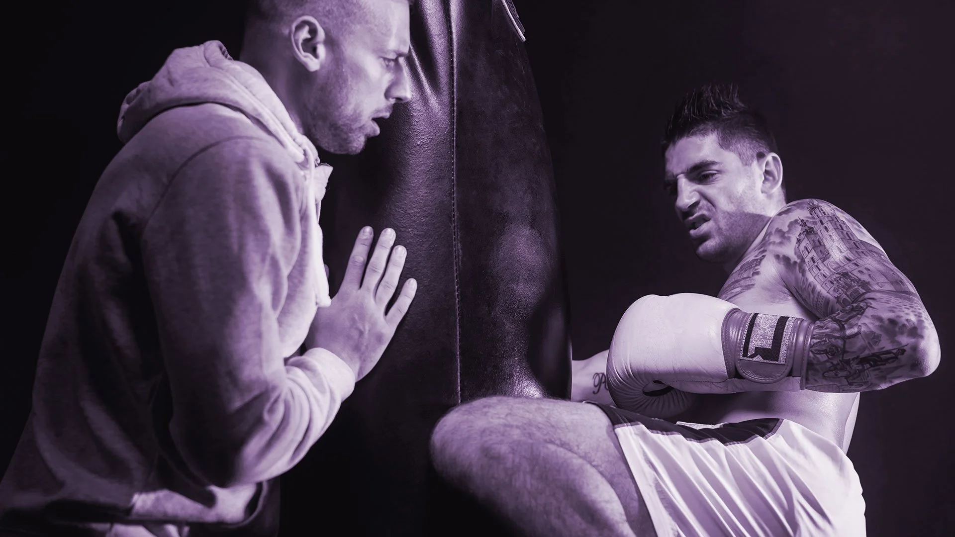 Un entrenador y un boxeador en un entrenamiento. El entrenador está apoyado en un saco de boxeo, mientras que el boxeador, con tatuajes en el brazo, usa guantes de boxeo y está recargado en la esquina del saco.