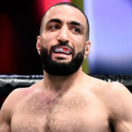 Hombre con barba, sin camiseta, con expresión de esfuerzo o incomodidad en un entorno de lucha libre o artes marciales.