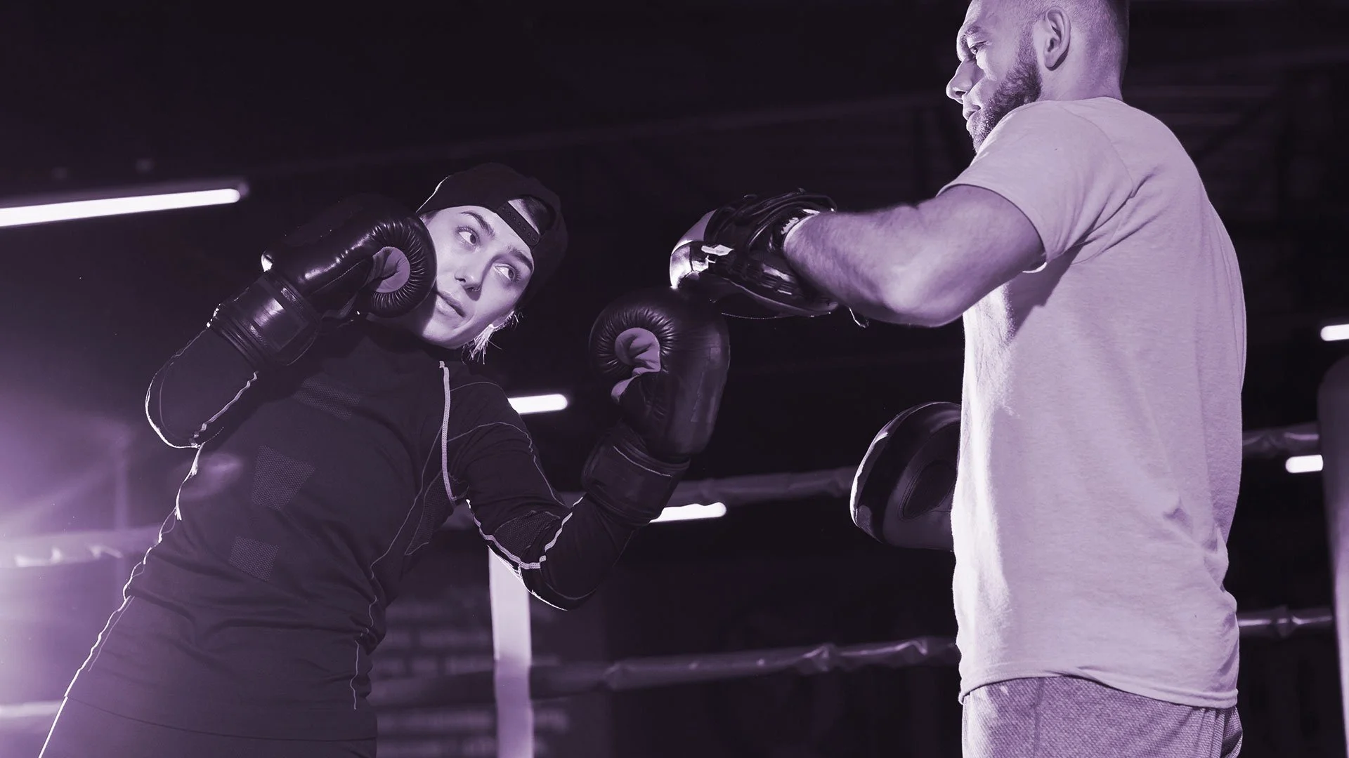 Una mujer y un hombre en un gimnasio entrenando boxeo. La mujer usa guantes de boxeo y protector en los oídos, y el hombre la está ayudando o entrenando.