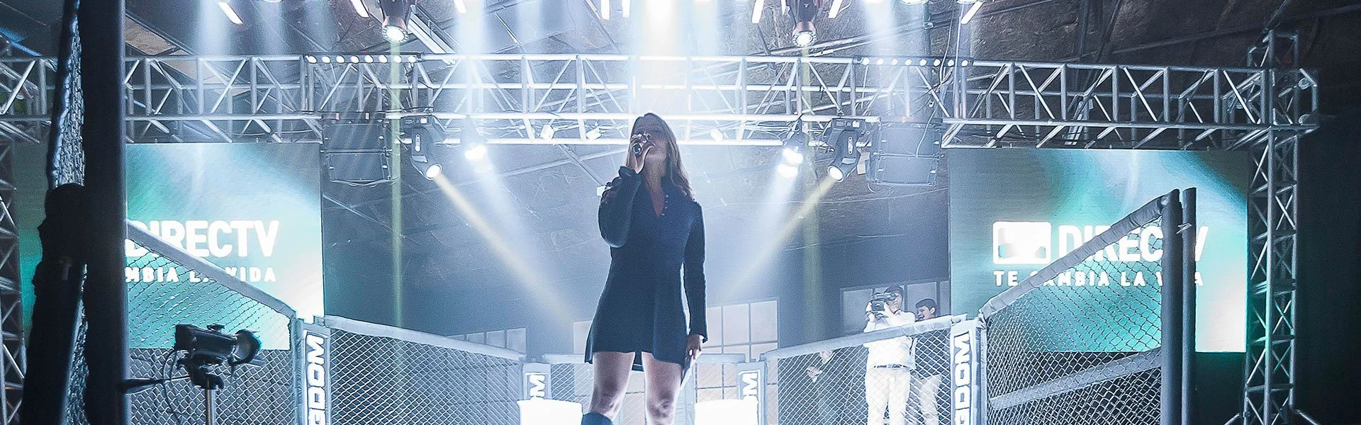 Mujer con vestido negro y camisa de mezclilla en la jaula de un evento de artes marciales mixtas, con pantallas que dicen 'DIRECTV TELEVISION LA' y iluminación teatral.