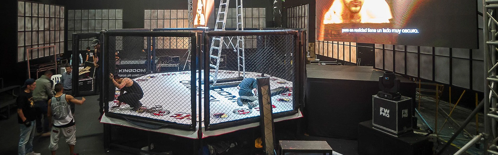 Dentro de una jaula de artes marciales en un escenario, con personas alrededor y un monolito grande con pantalla
