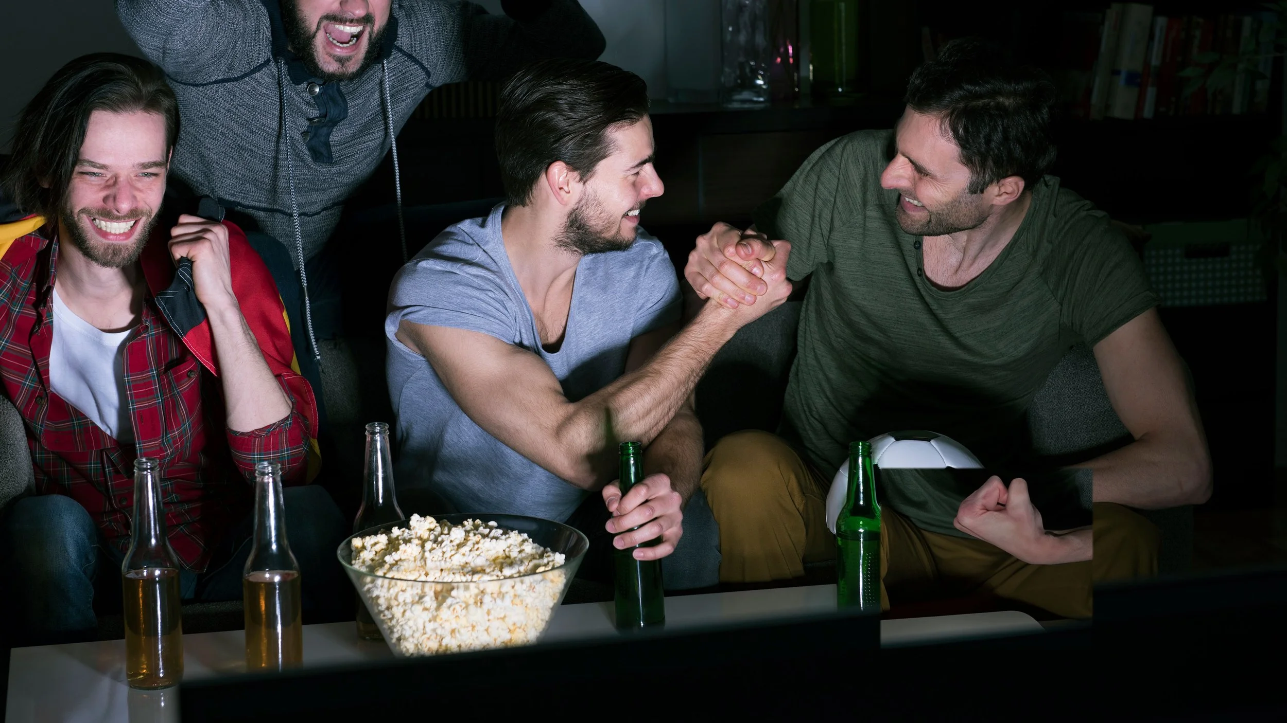 Grupo de amigos en una reunión, algunos sonriendo y saludándose con un apretón de manos, con botellas de bebidas y palomitas en la mesa.