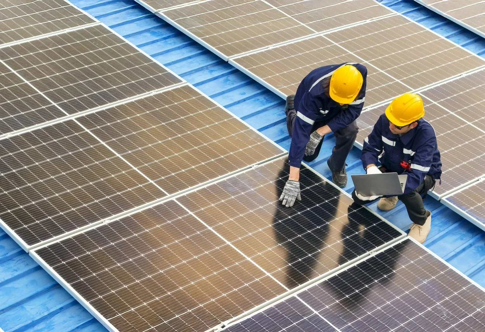 Two workers in yellow safety helmets and work uniforms installing or inspecting solar panels on a roof, one holding a laptop and the other adjusting a panel.