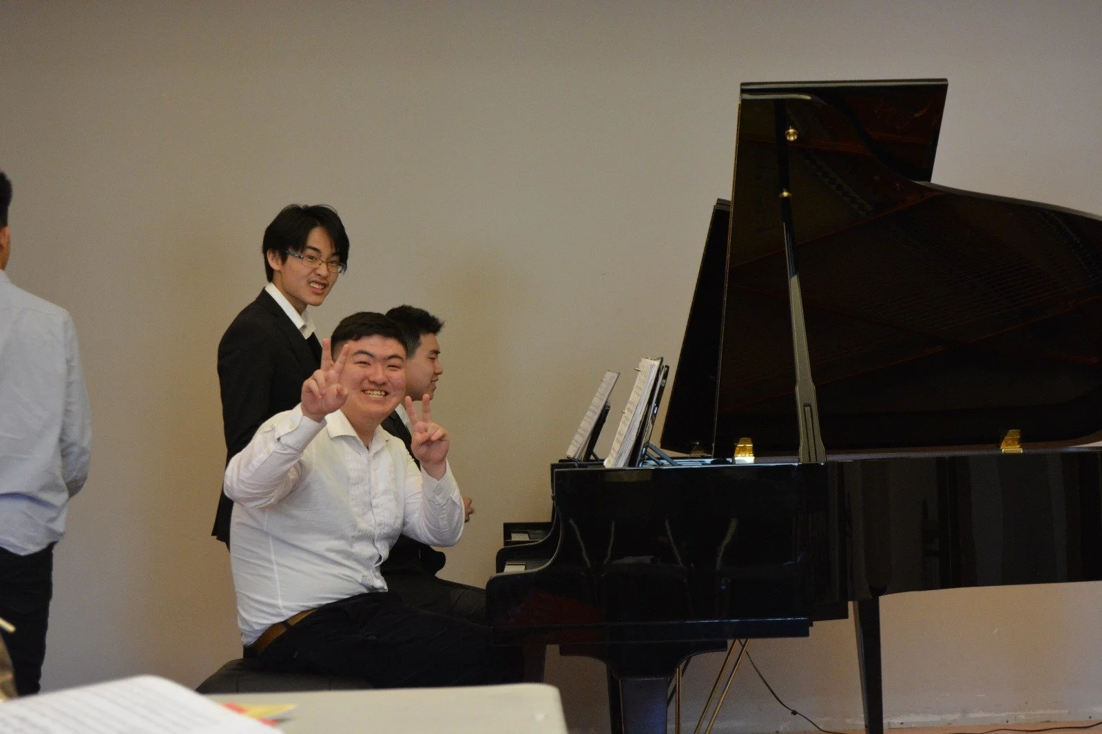 Young man smiling and making peace signs while sitting at a grand piano in a music room, with two other young men and a woman in formal attire standing nearby.
