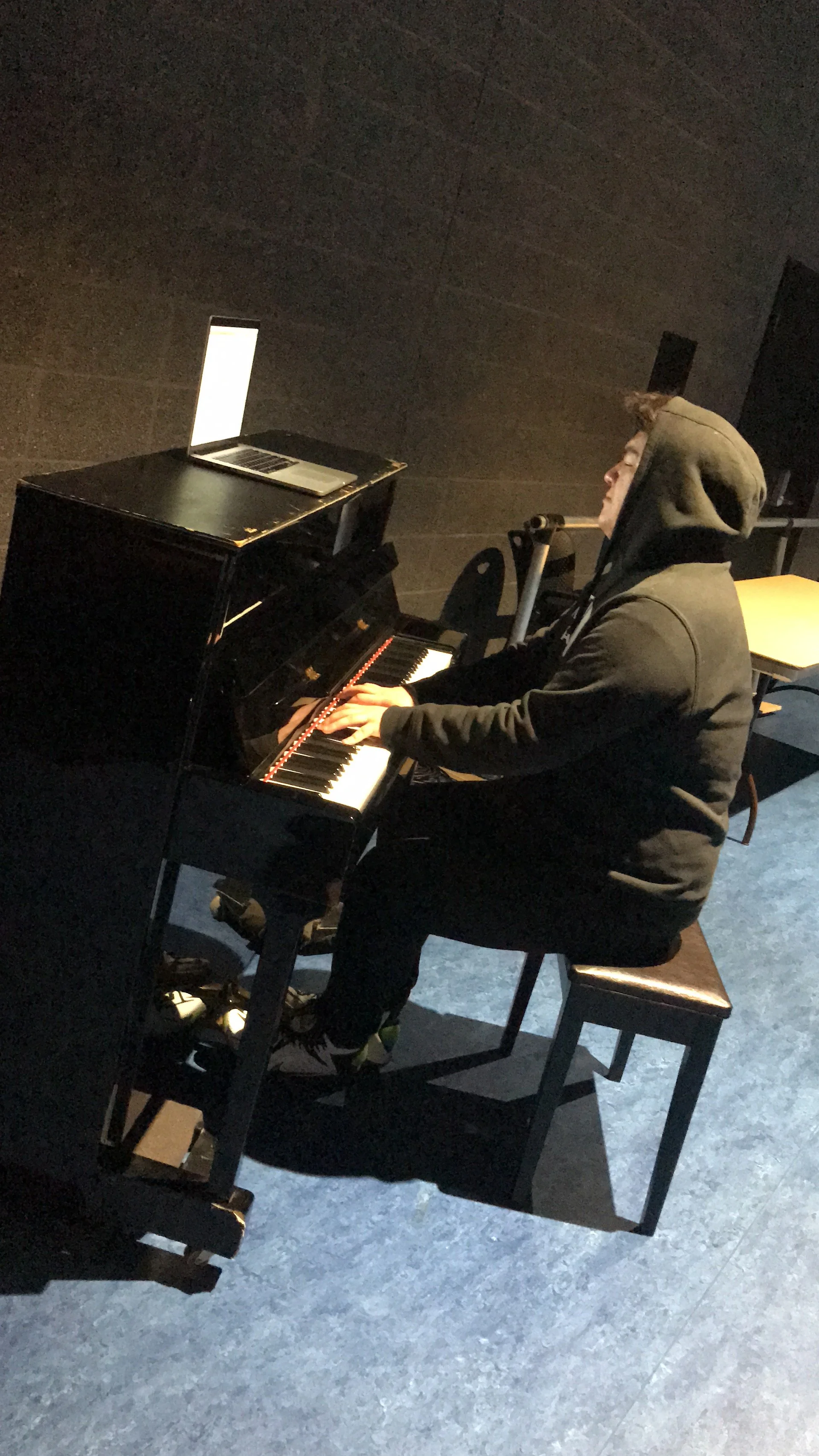 Person wearing a hoodie playing a black grand piano in a dimly lit room with a laptop on top of the piano.