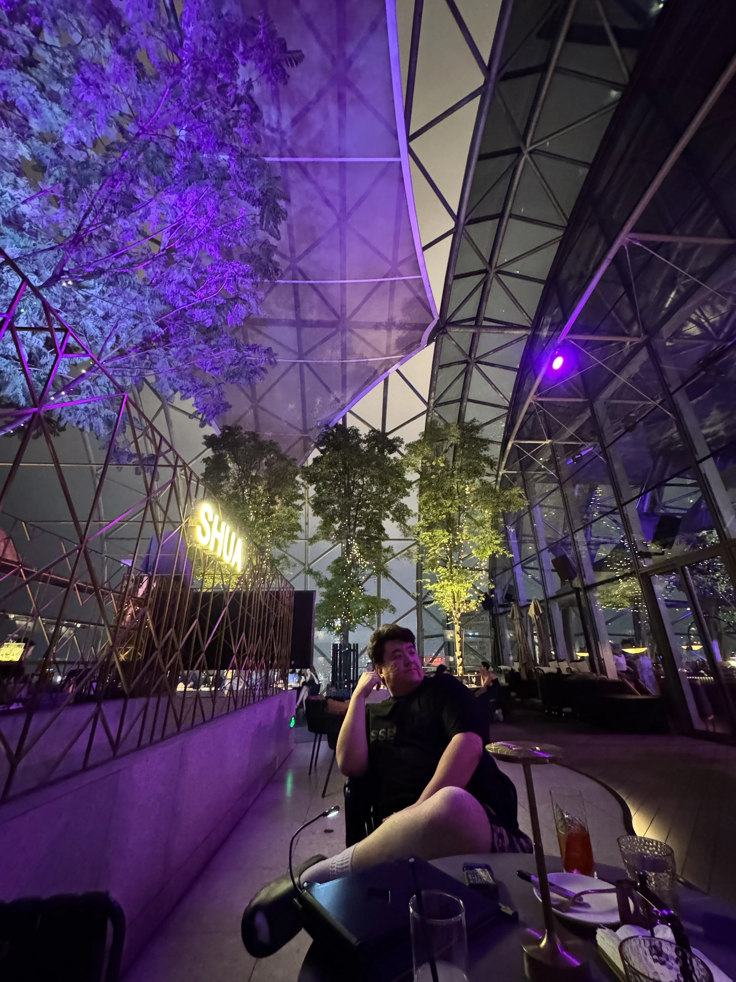 A person relaxing at a table in a modern indoor space with glass walls, illuminated trees, and colorful purple lighting, with a neon sign that reads 'SHU' in the background.