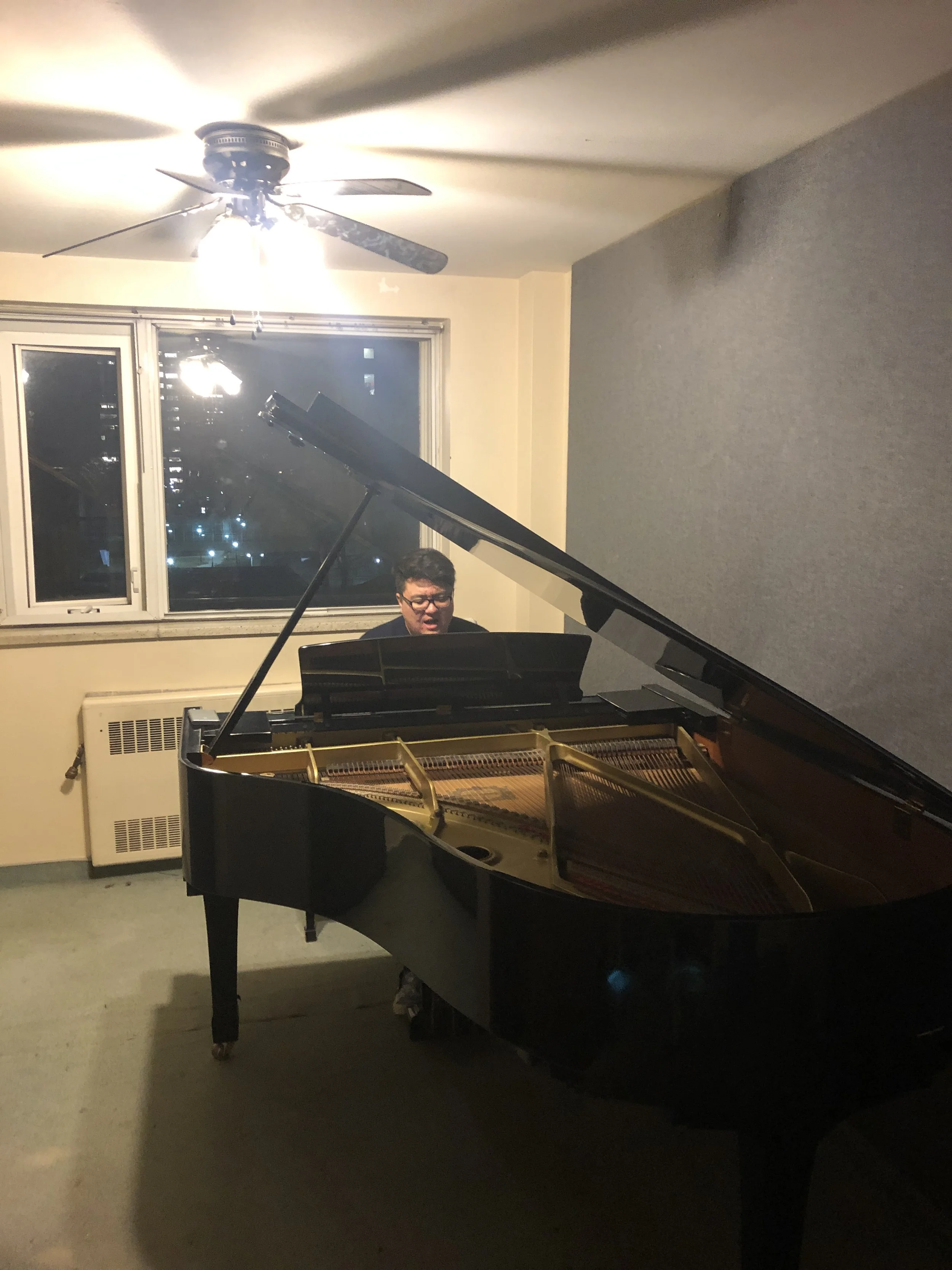 A person playing the piano in a room at night, with a ceiling fan and window showing city lights outside.