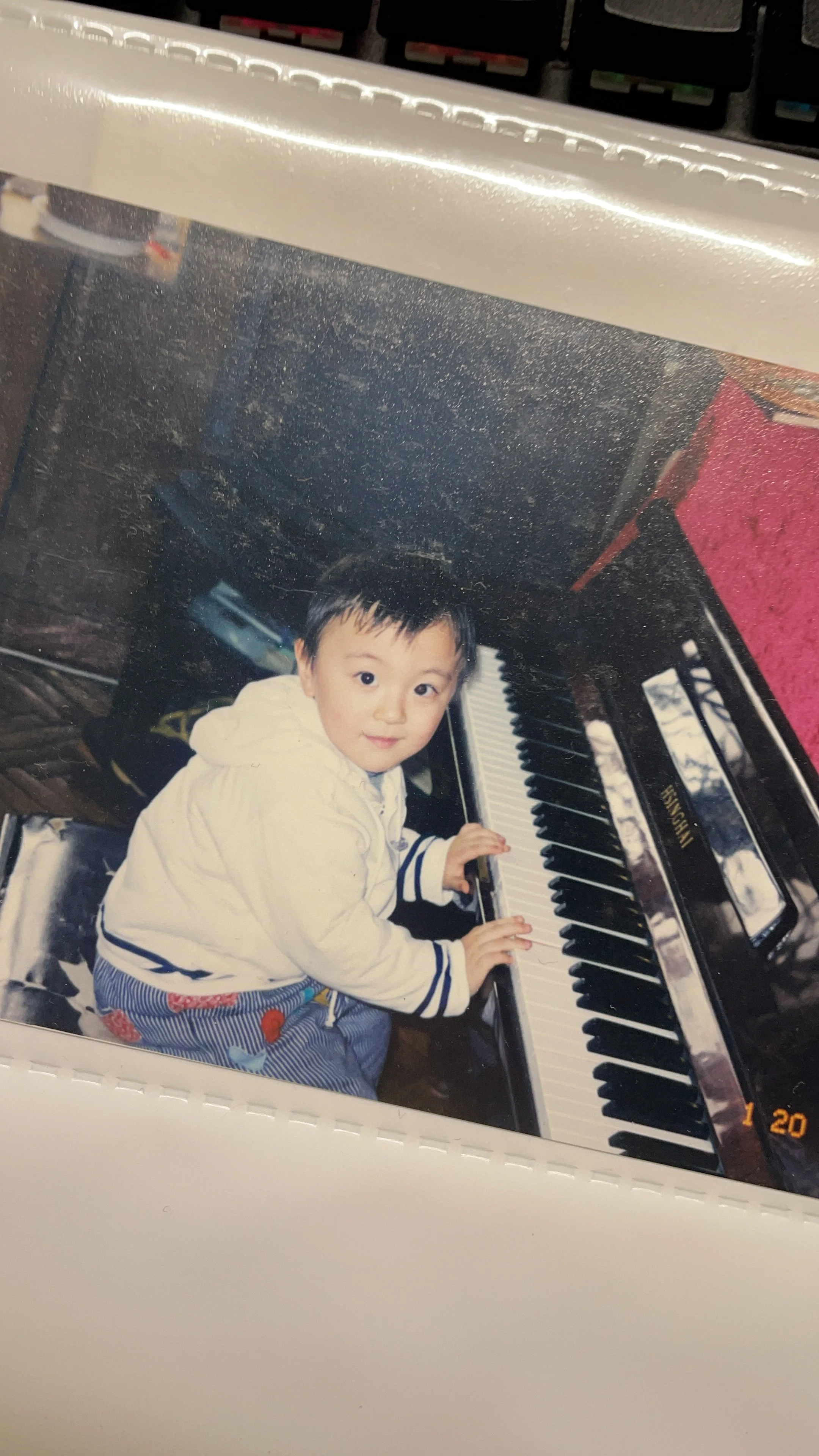 A young child with dark hair, wearing a white jacket and striped pants, sitting at a shiny black upright piano with the brand name 'Kawai', looking up at the camera with a slight smile.