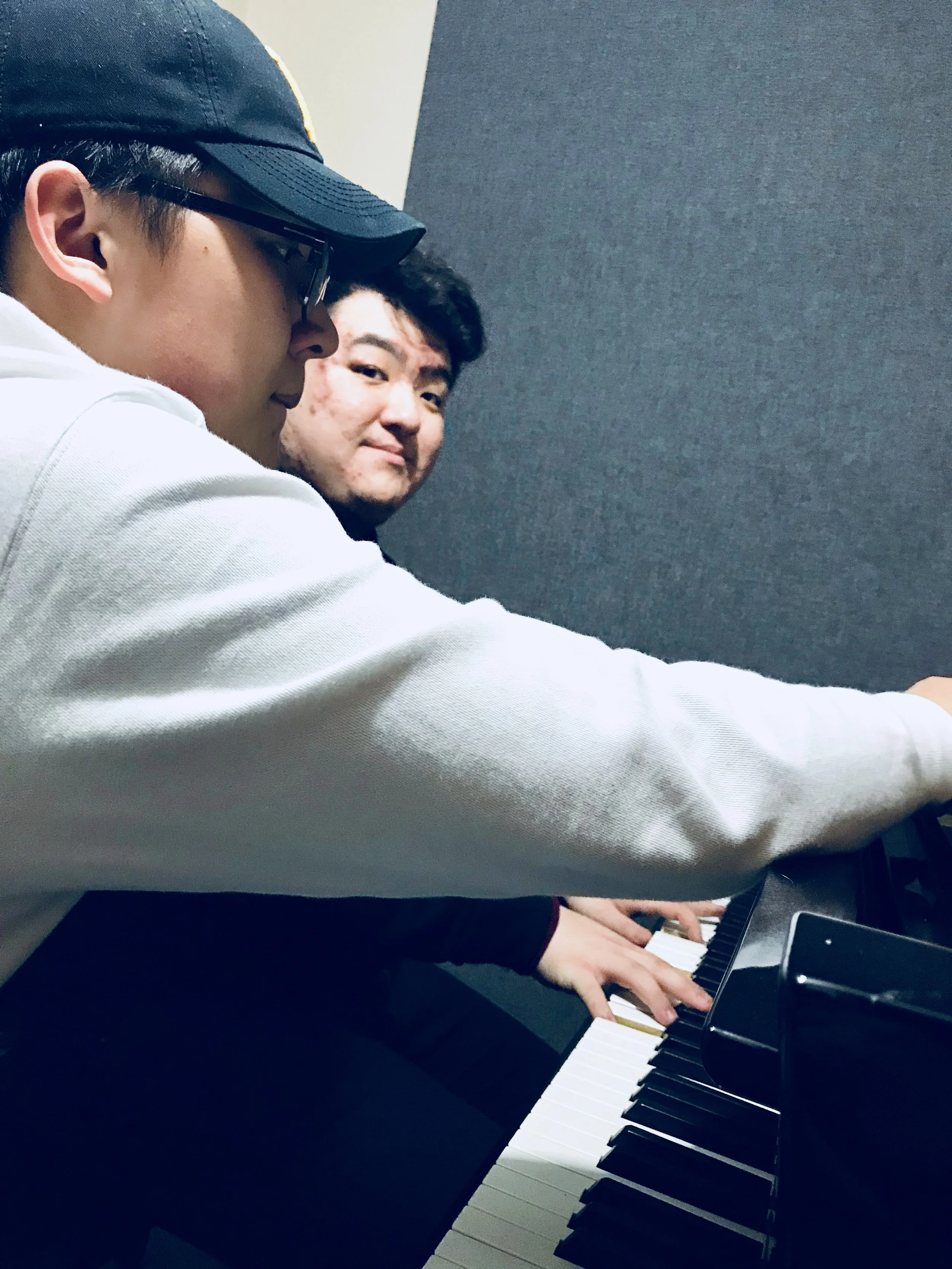 Two young men playing a black grand piano; one wearing a baseball cap and glasses, the other with curly hair, both focused on the keys in a music room.