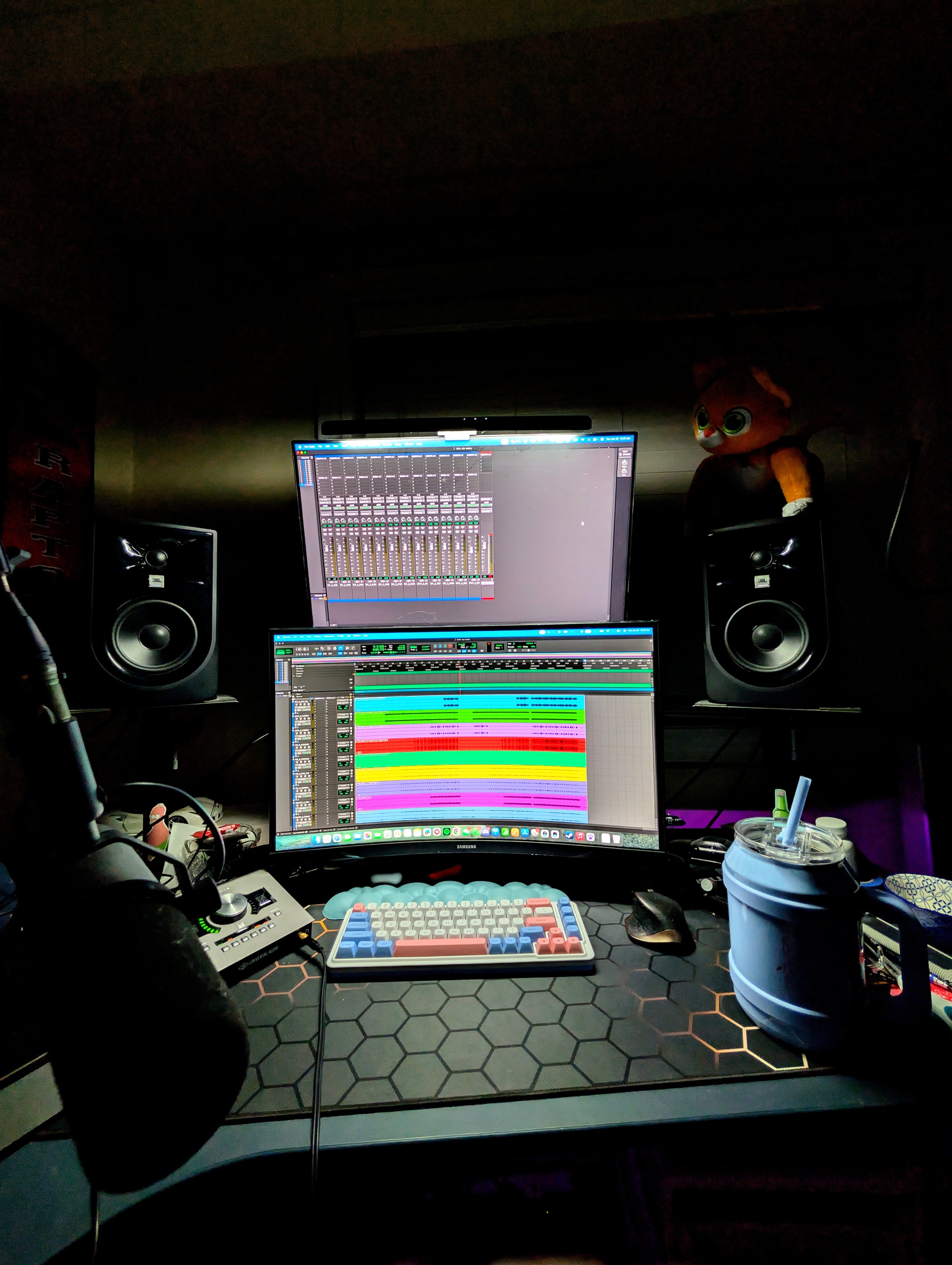 A music production workspace with two monitors displaying digital audio workstation software, a keyboard, a mouse, studio speakers, a microphone, and a stuffed animal on the right speaker. There is a drink container on the right side of the desk.