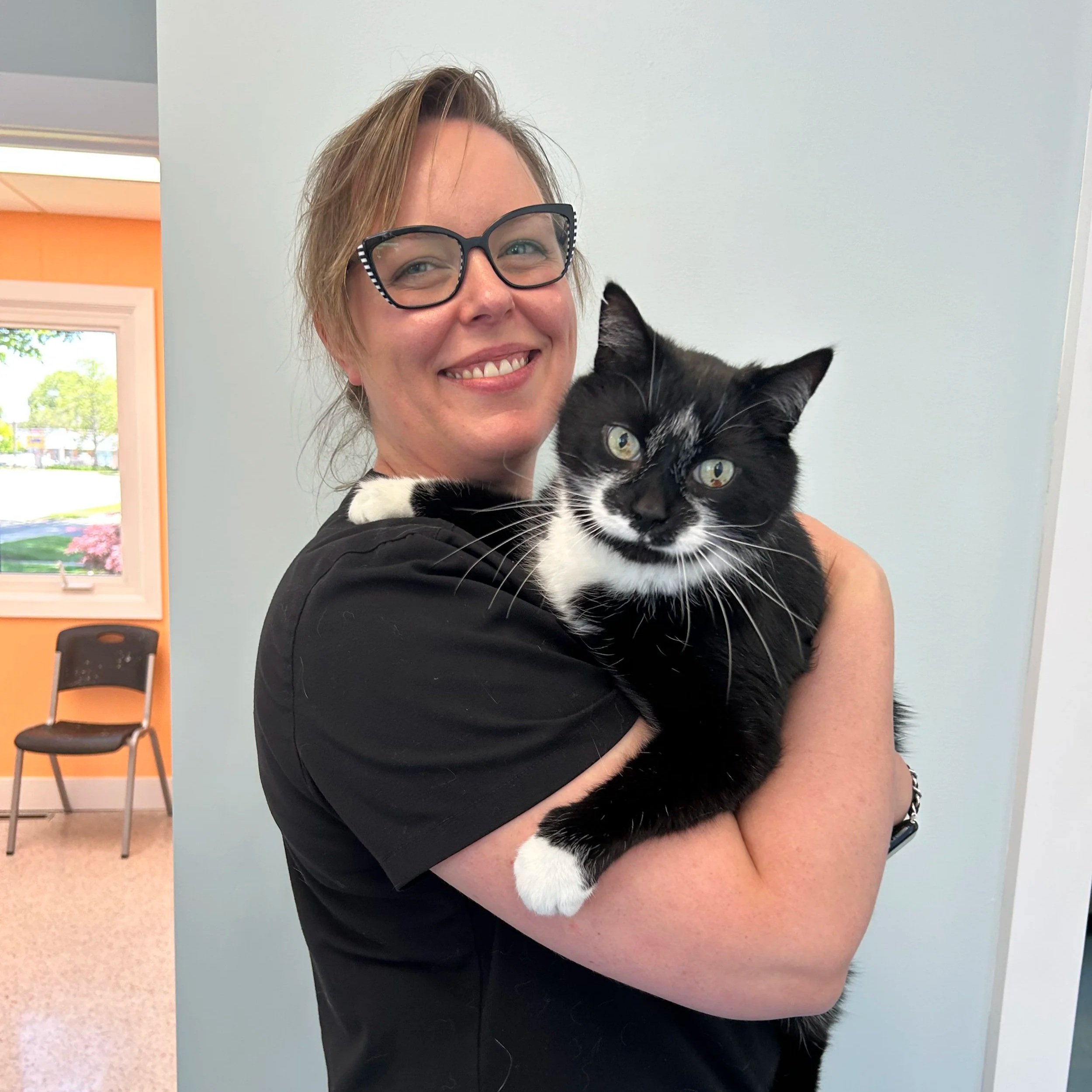 A woman with short hair and glasses is holding a black and white cat. The woman is smiling and the background shows an orange wall with a window and some chairs.