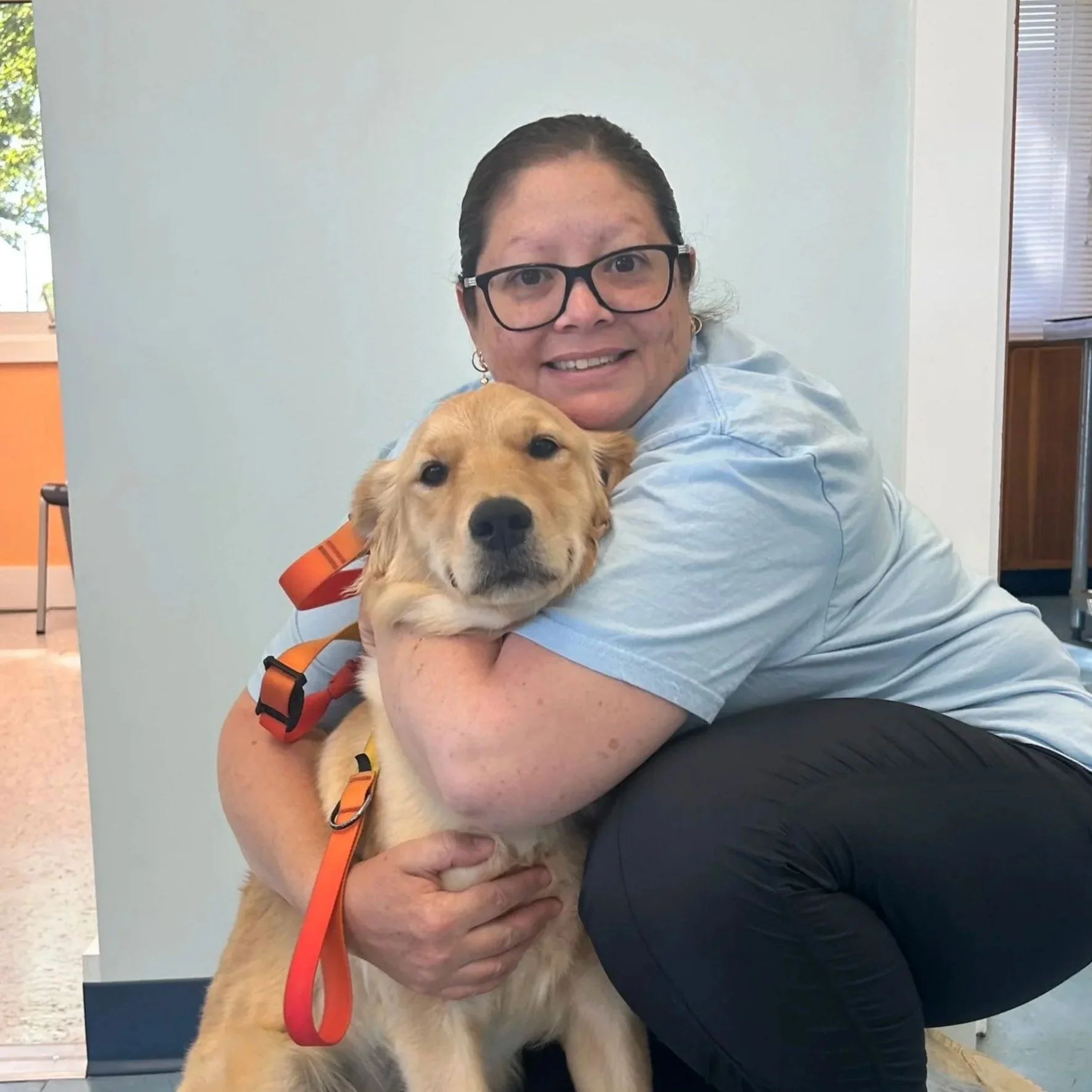 A woman with glasses hugging a golden retriever puppy, both smiling, indoors with white walls and windows in the background.