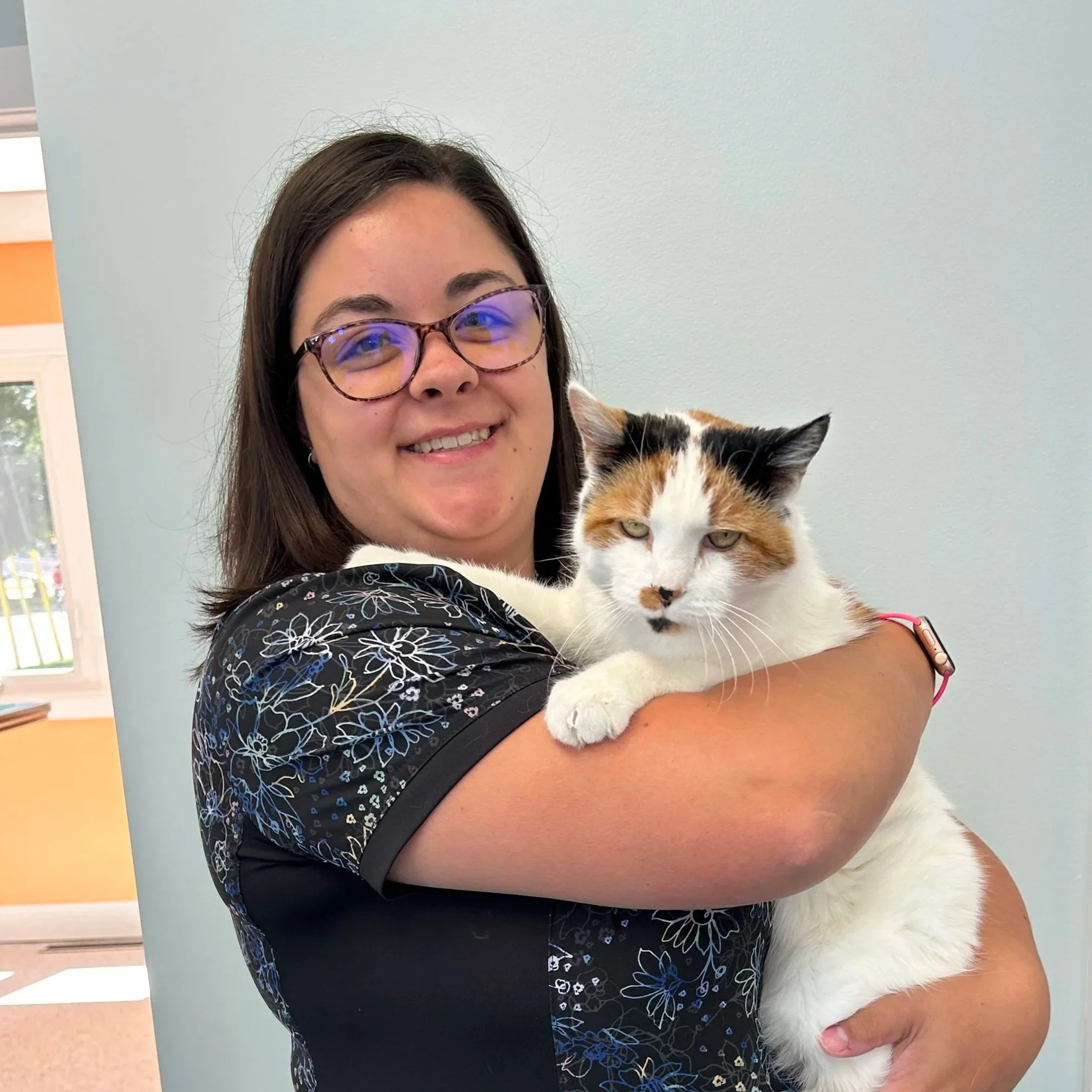 A woman with long brown hair wearing glasses, smiling, holding a calico cat with white, orange, and black fur.