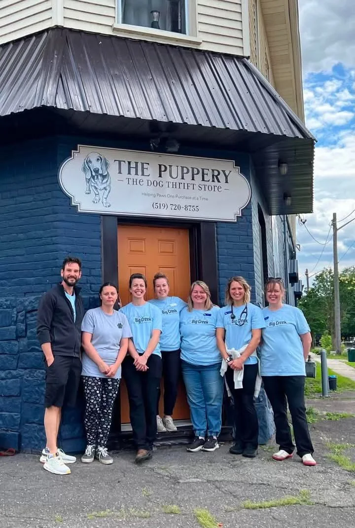 Group of seven people standing in front of a building with a sign that reads 'The Puppery - The Dog Thrift Store.' Six women and one man, all smiling, with the women wearing light blue T-shirts that say 'Big Creek.'
