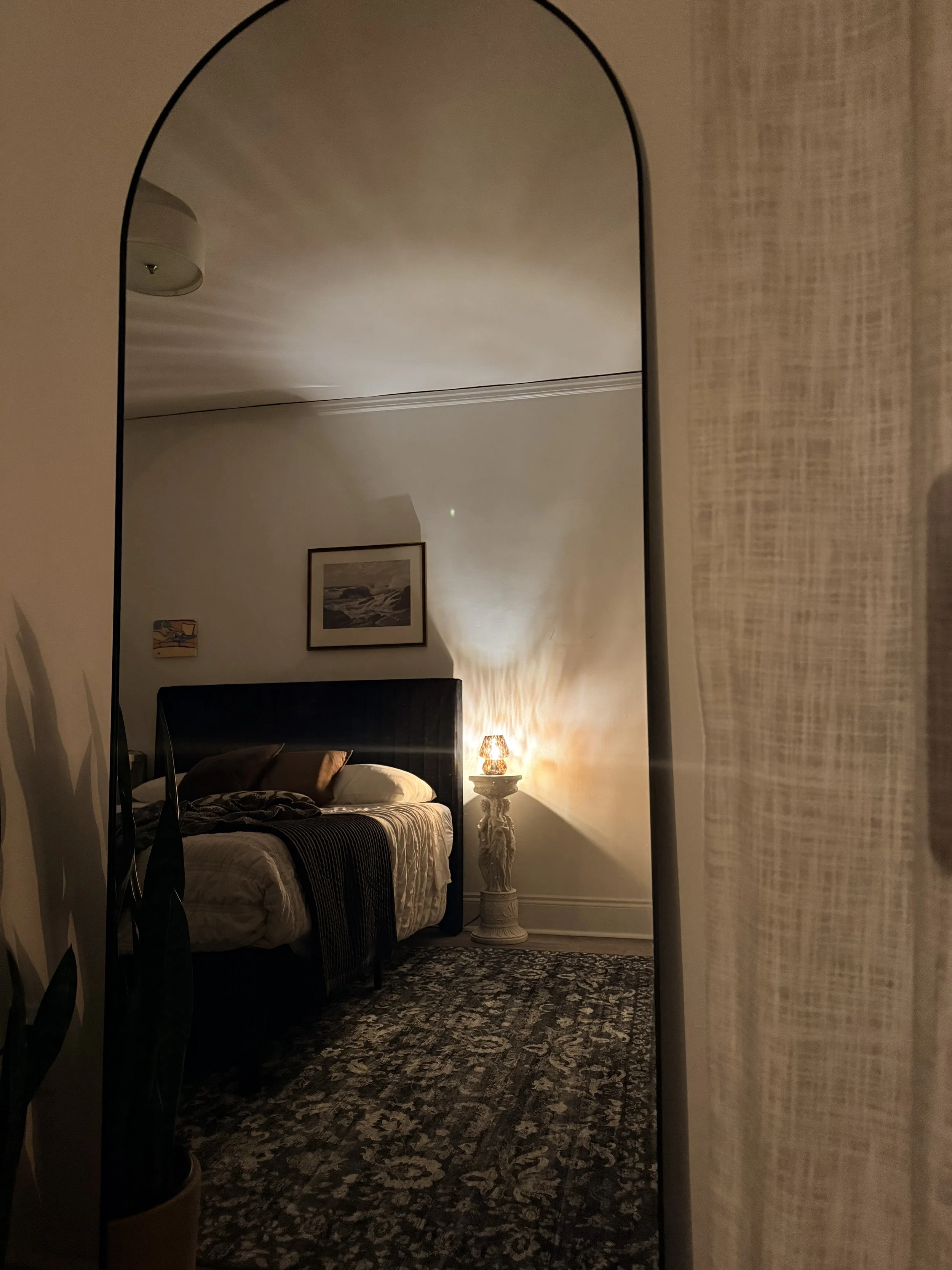 Arched floor mirror reflecting a warmly lit bedroom with a bed, framed artwork, table lamp, and patterned rug