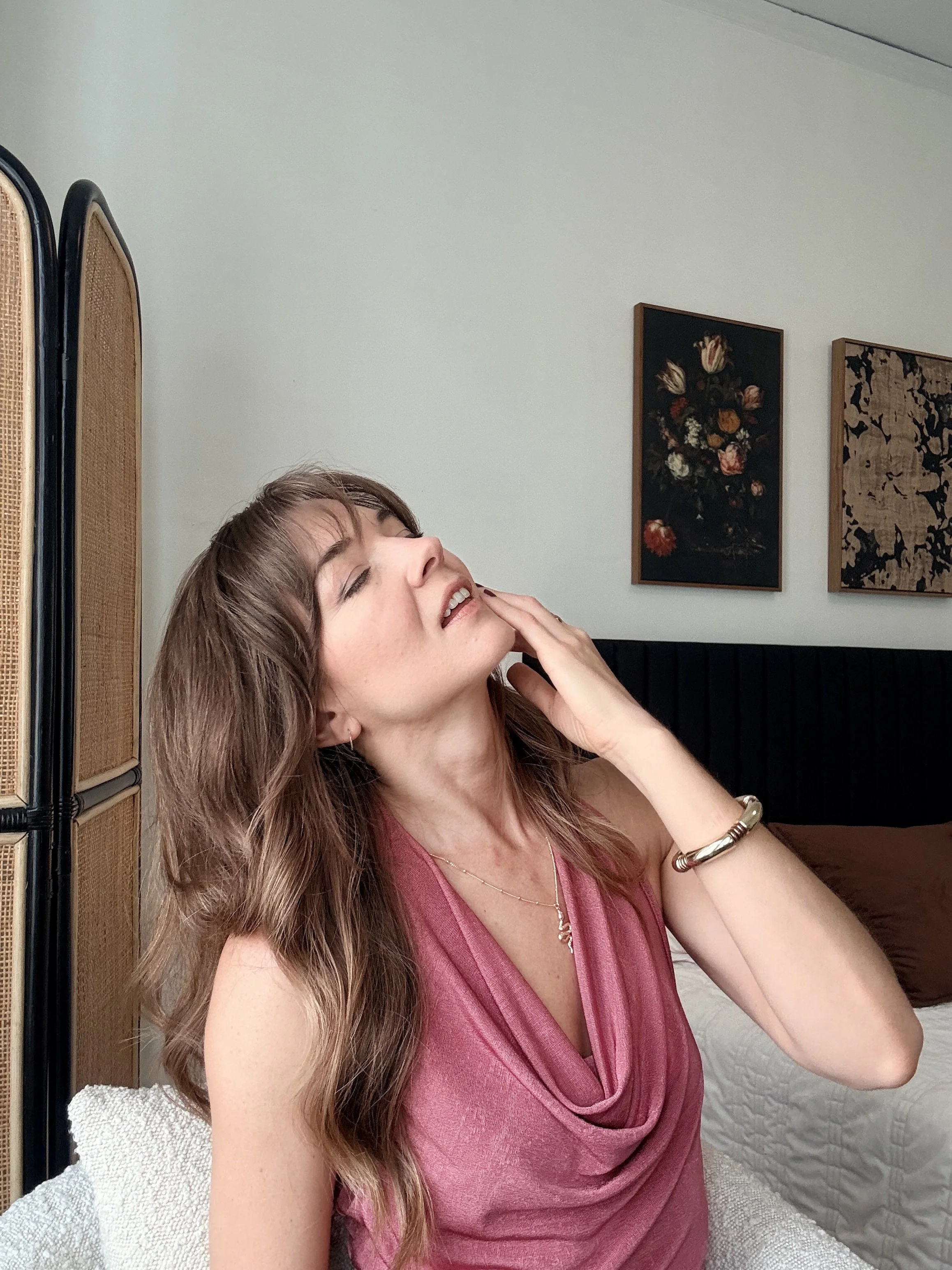Woman in a pink top smiling and touching her cheek while seated indoors, with framed floral artwork behind her.