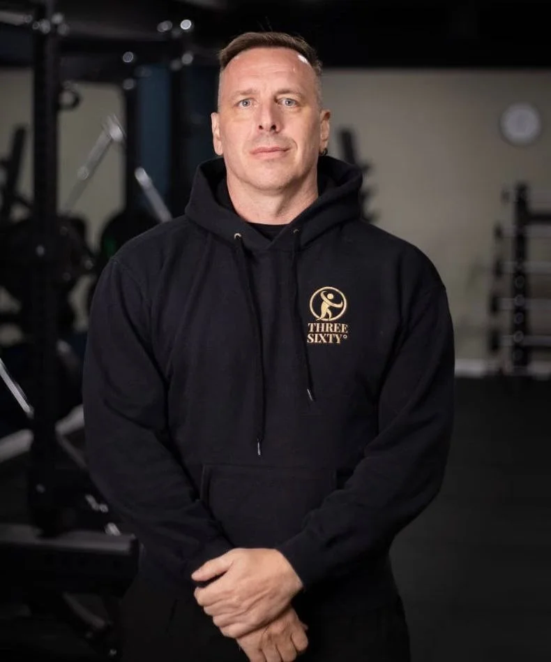 A man wearing a black hoodie with a gold logo and text that says 'THREE SIXTY' standing in a gym with weights in the background.
