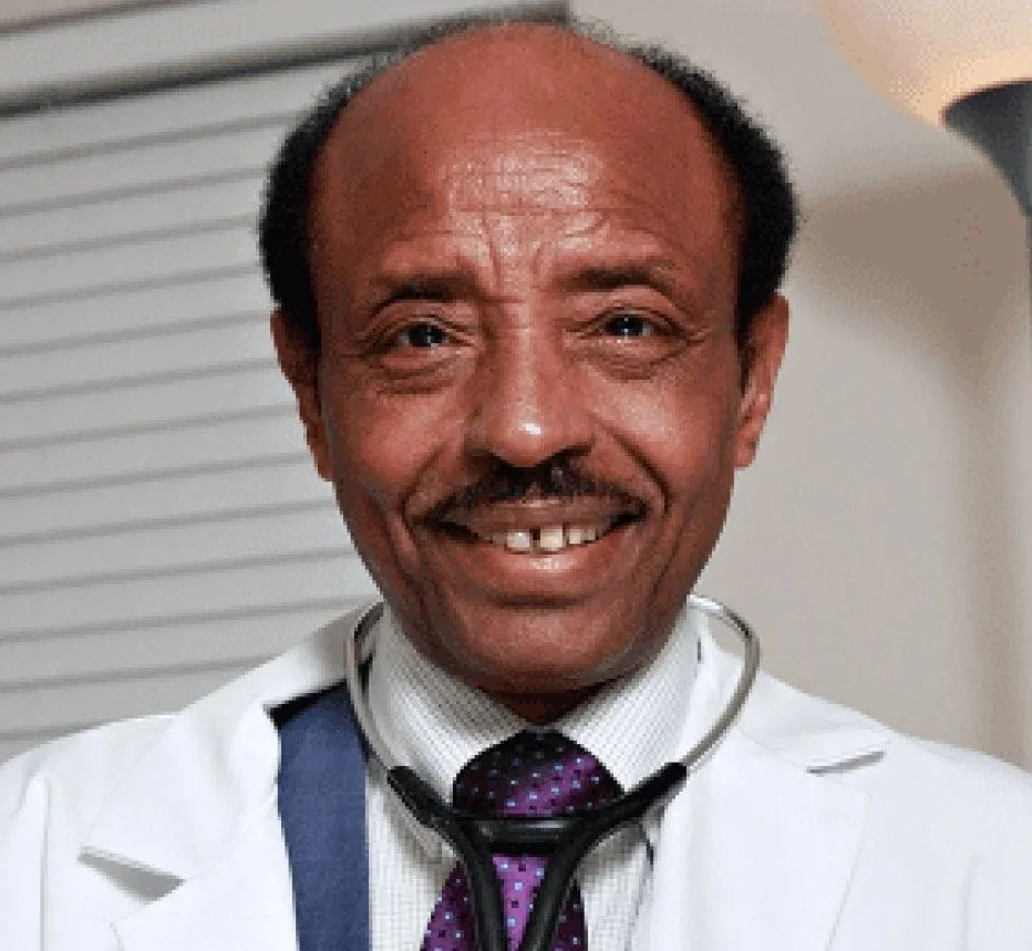 A smiling male doctor with a mustache, wearing a white coat, a purple tie, and a stethoscope around his neck, indoors with a white wall and window blinds in the background.