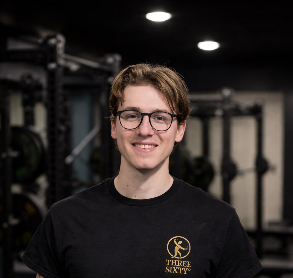 Young man with glasses smiling in a gym, wearing a black T-shirt with a logo and text, with gym equipment in the background.