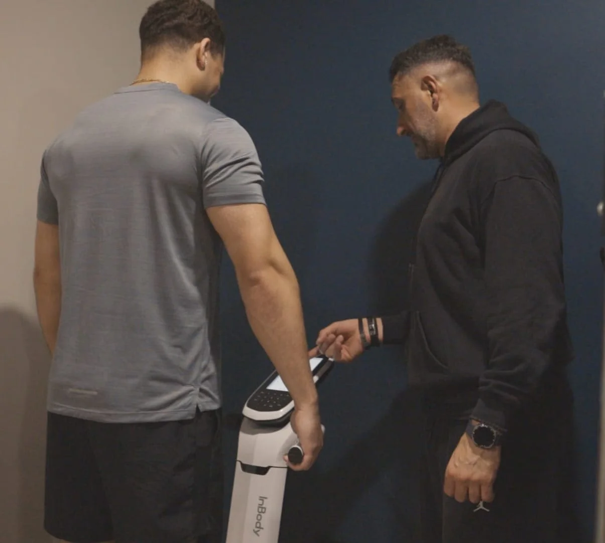 Two men are standing and looking at a medical or fitness device with a screen, with a dark blue wall in the background.