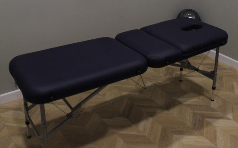 A black massage or treatment table with adjustable sections, set up in a room with wooden flooring and a plain wall.