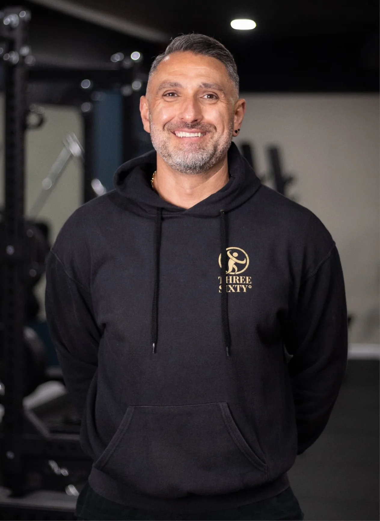 A smiling man with short gray hair and a beard wearing a black hoodie with a logo on the chest, standing in a gym with exercise equipment in the background.
