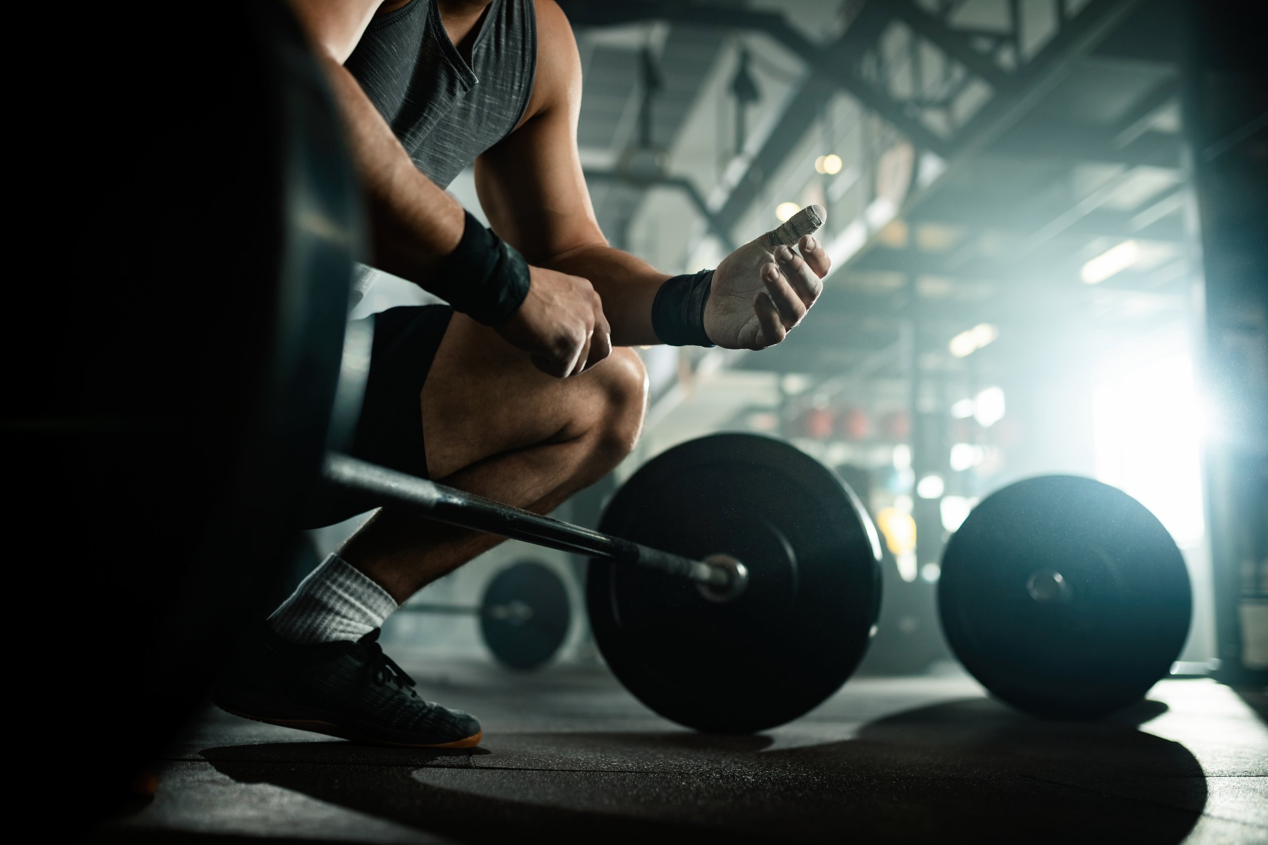 A person in athletic attire is sitting on a bench in a gym, holding a weight in their hand. Dumbbells are visible on the floor nearby, and the gym has an industrial-style interior with bright light coming from a window or door.