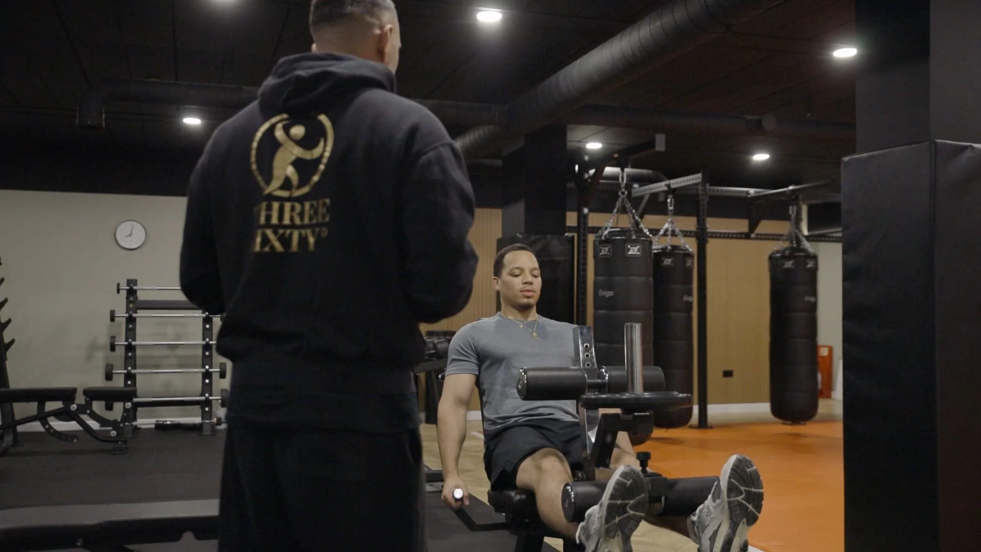 Two men in a gym, one sitting on a rowing machine and the other standing next to him. The man on the rowing machine has short hair, a grey T-shirt, and shorts. The other man has short hair, a hoodie with a logo on the back, and dark pants. The gym has punching bags in the background and a clock on the wall.