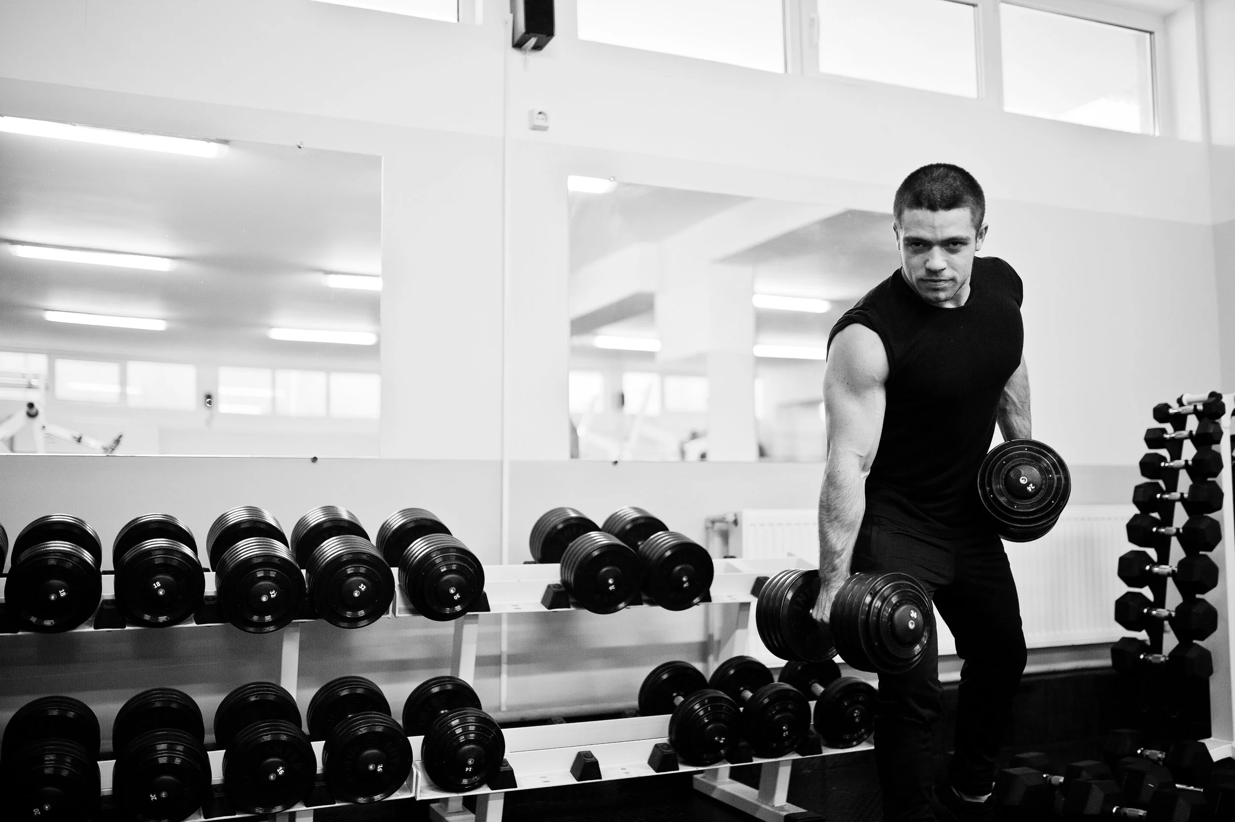 A man working out with dumbbells in a gym, standing beside a rack of black dumbbells, in front of a mirror, with bright indoor lighting.