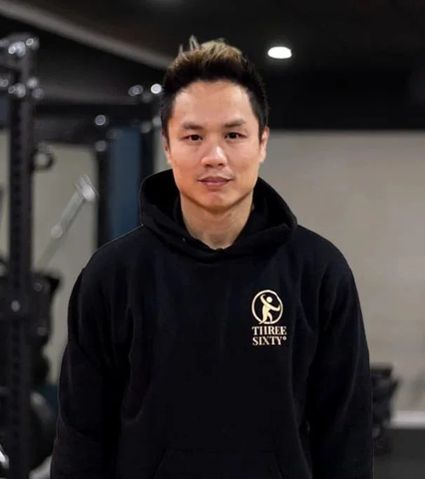 A young man with short dark hair wearing a black hoodie with a logo on the front that reads 'THREE SIXTY' and depicts a stylized figure doing a yoga pose, standing in a gym or fitness center.