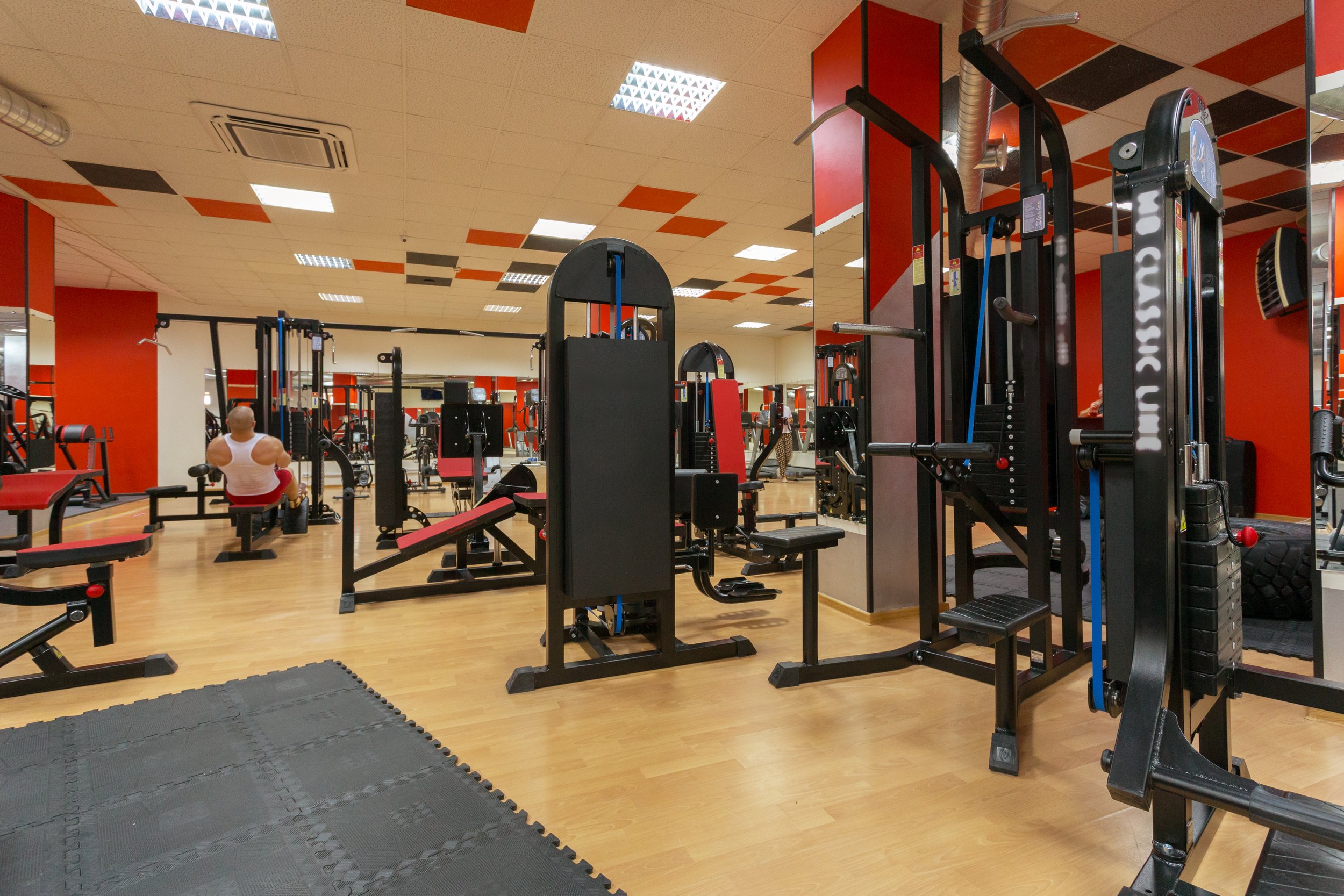 An empty gym with black and red workout equipment, including weight machines and benches, on a wooden floor. There are a few people working out in the background, and the ceiling has white tiles with orange and black patterns.