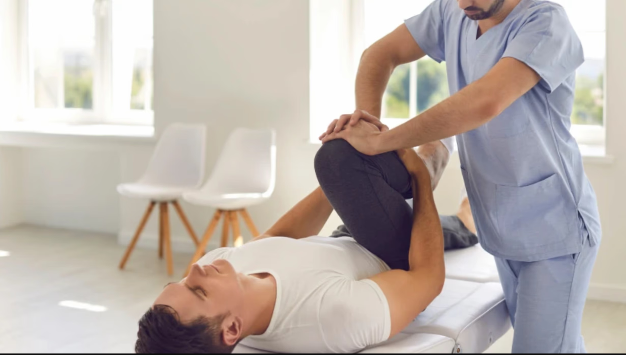 A healthcare professional performing physical therapy or chiropractic care on a male patient lying on a treatment table, lifting his leg while holding it.