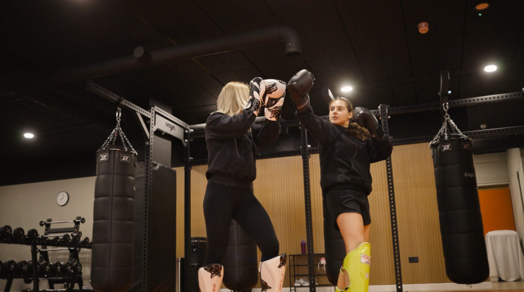 Two women practicing boxing in a gym, one teaching the other, with punching bags hanging in the background.