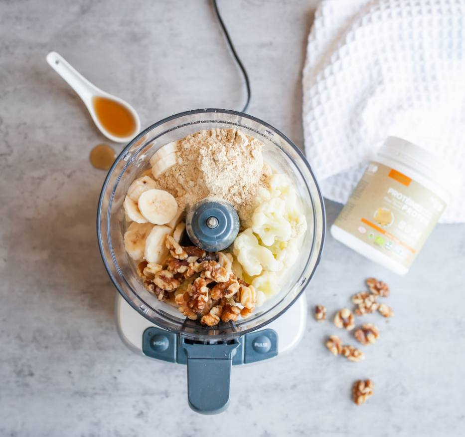 A food processor containing sliced bananas, walnuts, mashed cauliflower, protein powder, and flour on a gray countertop. Nearby are a small spoon with honey, some loose walnuts, a container of protein powder, and a white textured towel.