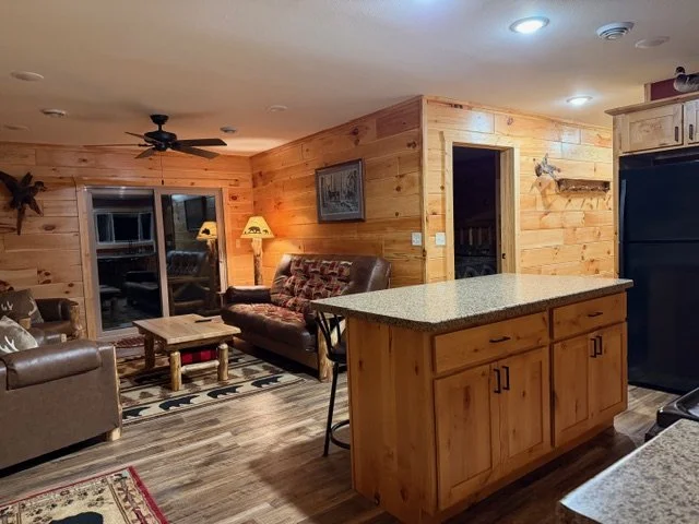 A rustic kitchen with wooden cabinets and a kitchen island with a granite countertop, black refrigerator, stove, and small appliances on the counter, in a cabin-style interior.