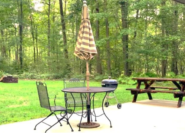 Patio area with a round metal table, two metal chairs, a closed striped patio umbrella, a charcoal grill, and a wooden picnic table, surrounded by trees and grass.