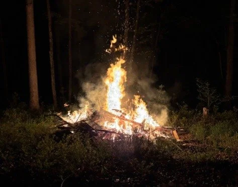 Campfire with flames and sparks rising into the night sky in a wooded area.