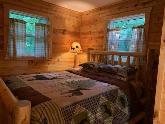 A cozy bedroom with wooden walls, a large bed with a patterned quilt and pillows, two windows with checkered curtains, and a bedside lamp with a nature scene on shade.