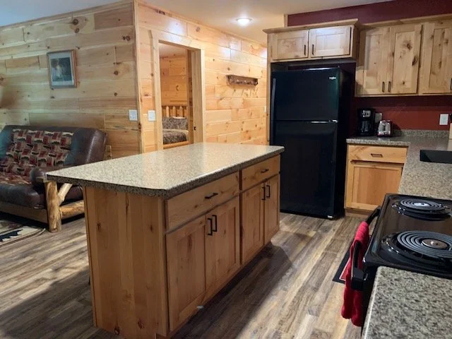 Kitchen with wooden cabinets, black appliances, and a granite countertop island, next to a living area with a patterned sofa.
