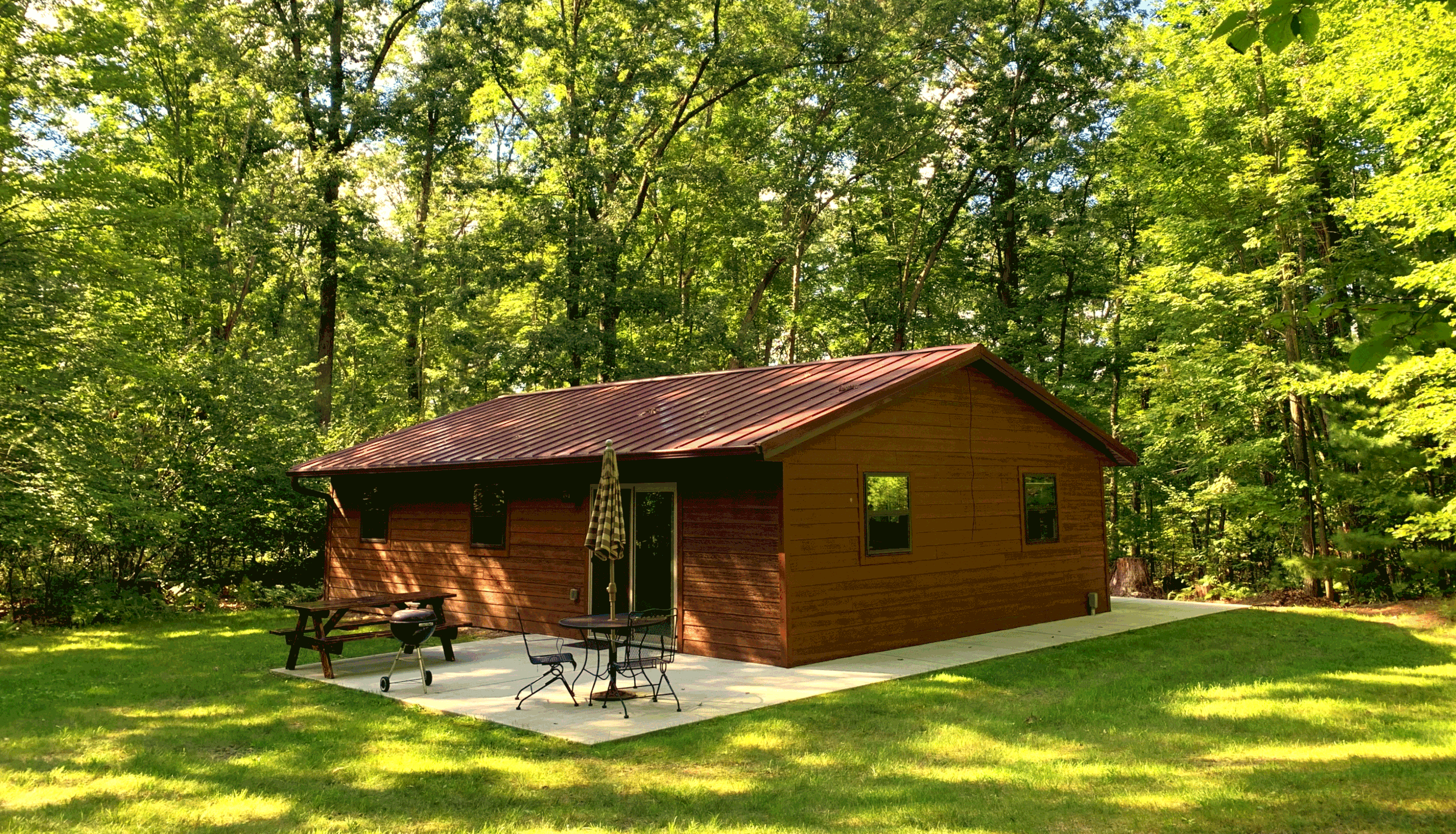 A small wooden house with a red metal roof in a lush wooded area with tall green trees. There is a concrete patio with outdoor furniture, including a picnic table, chairs, an umbrella, and a barbecue grill.