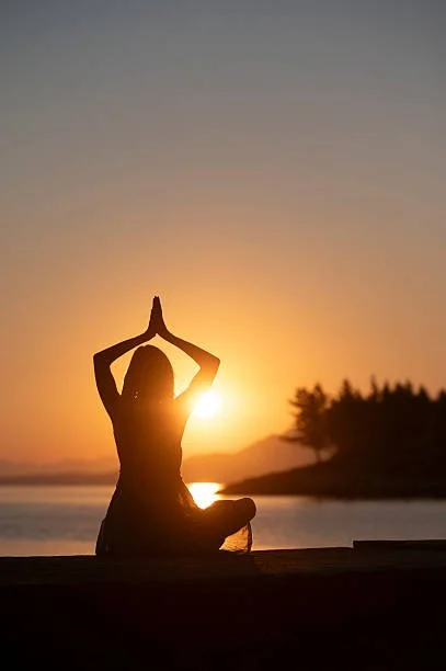Silhouette of a person practicing yoga in a seated meditation pose with hands in prayer position at sunset by a body of water, with trees and mountains in the background.
