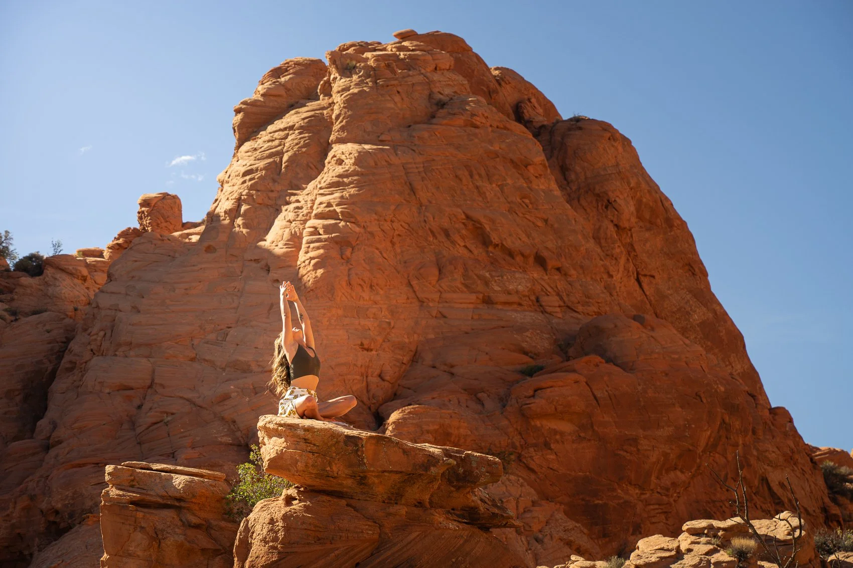 A woman practicing yoga or meditation in a headstand pose on a rocky outcrop in a desert landscape with red rock formations and a clear blue sky.