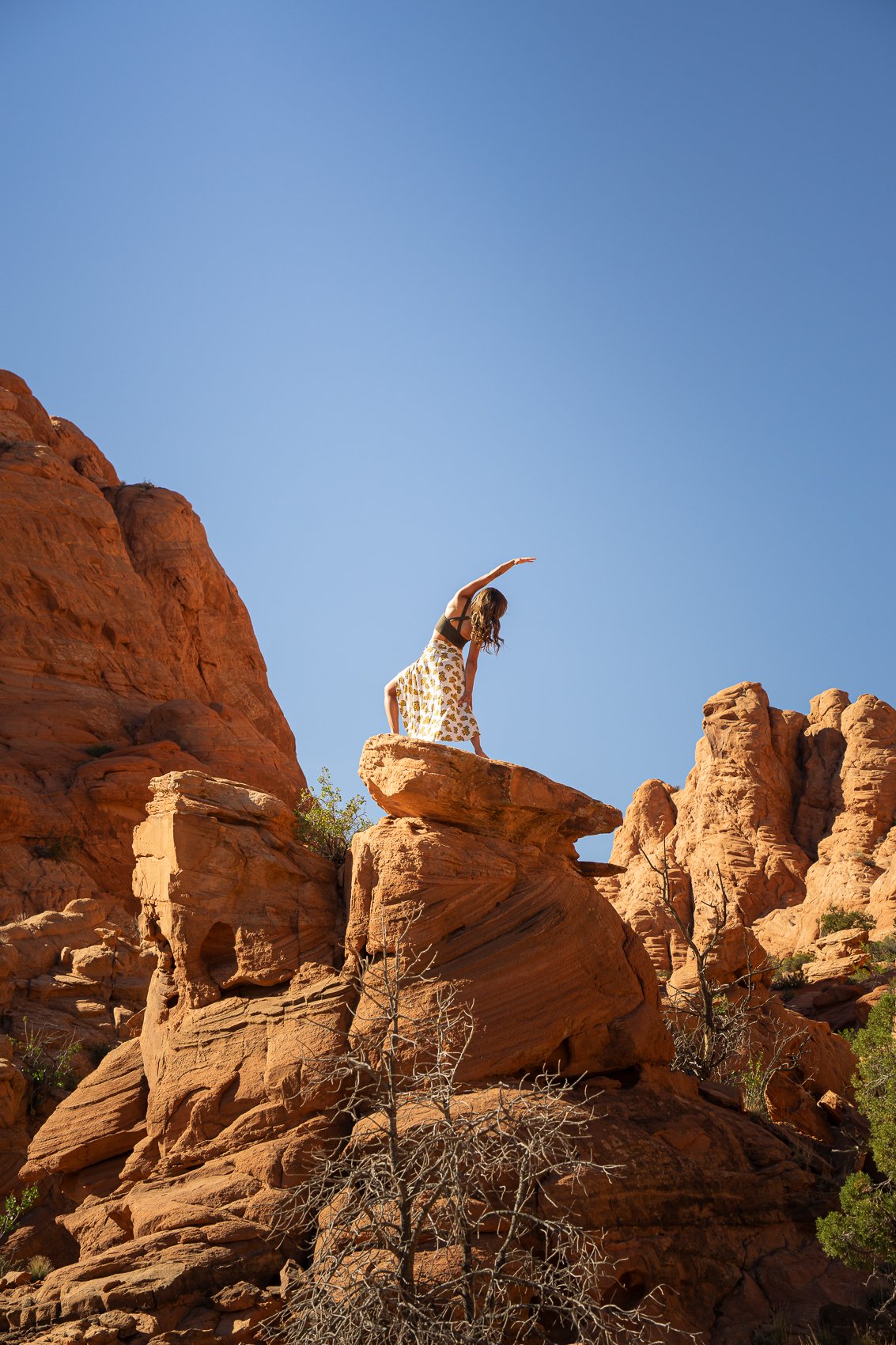 Woman in black top and patterned skirt balancing on a rock formation in a desert landscape with red rocks and a clear blue sky