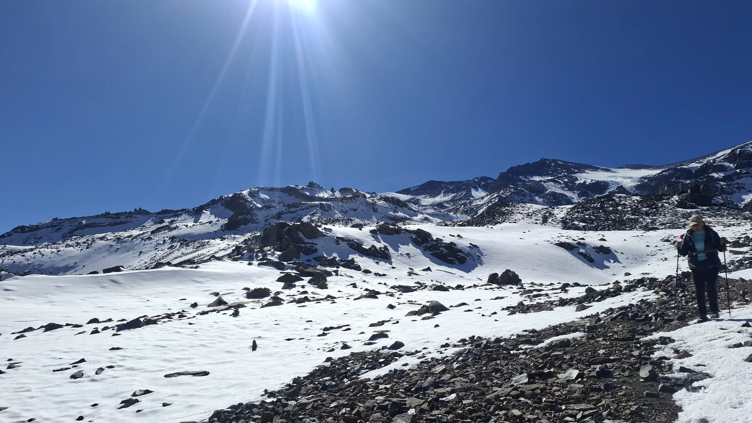 Persona haciendo senderismo en un paisaje de montaña nevada, con rocas y cielo despejado.