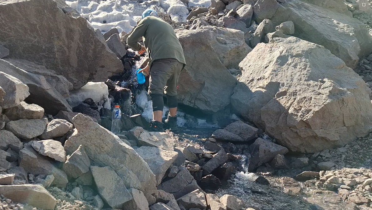 Persona que recolecta agua en un arroyo rodeado de rocas.